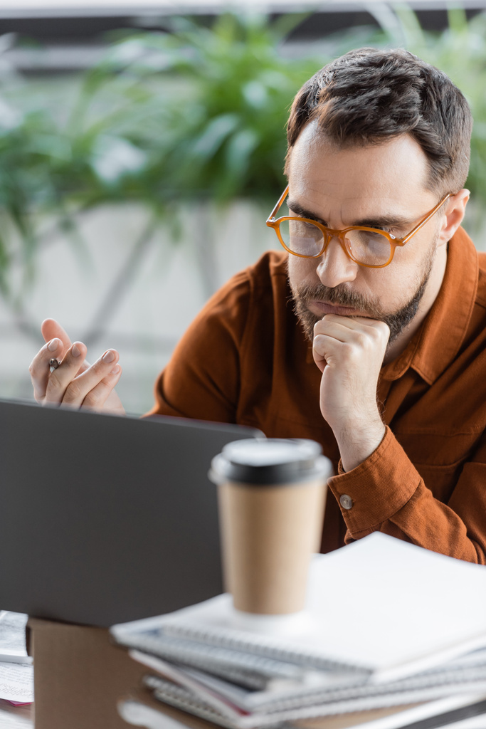 fáradt és figyelmes üzletember elegáns szemüveg mellett ül laptop kartondobozon, halom notebook és kávé menni, miközben megoldja a problémát elmosódott előtérben az irodában - Fotó, kép