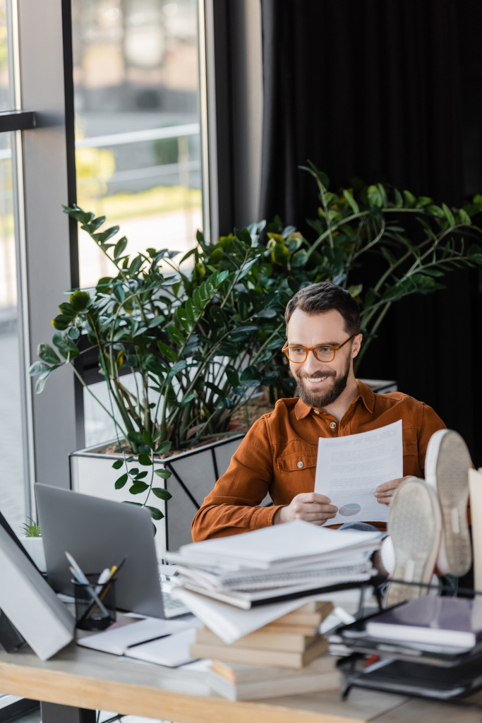 харизматичный и счастливый бизнесмен в очках держит документ, сидя с ногами на рабочем столе рядом с ноутбуком, кучей ноутбуков и декоративных растений в офисе - Фото, изображение