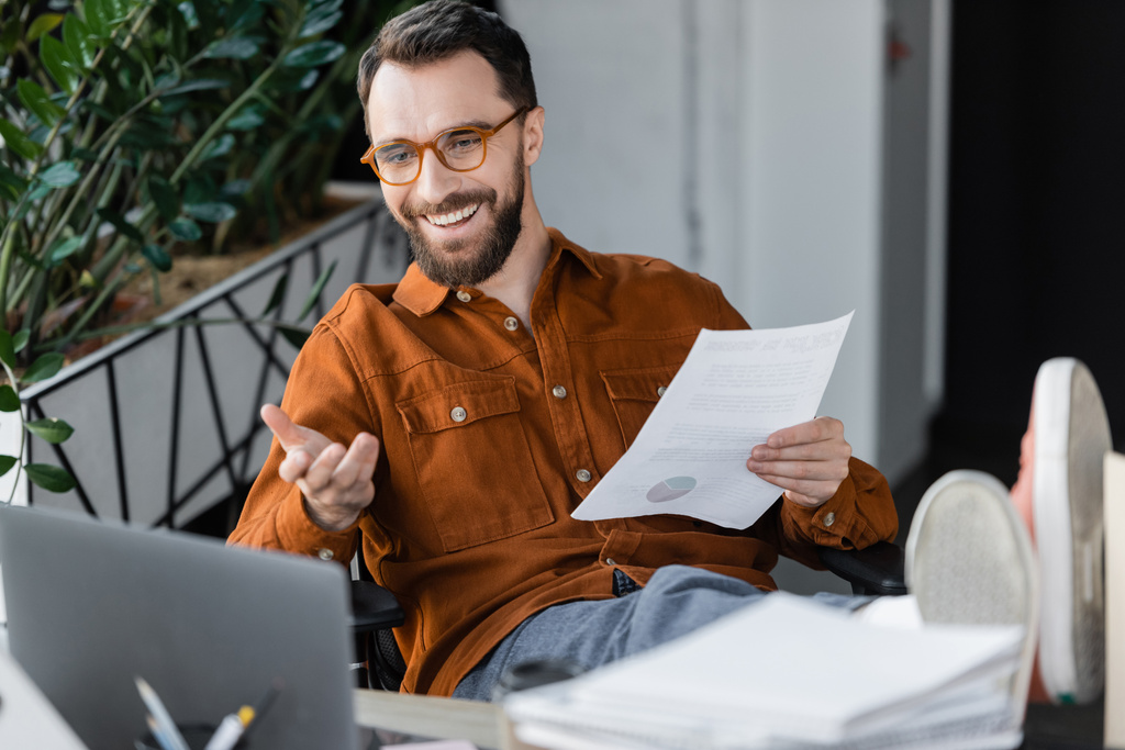 веселый бизнесмен в модных очках и футболках держит документ, сидя с ногами на столе и указывая рукой во время видеозвонка на ноутбуке рядом с бумагами на размытом переднем плане - Фото, изображение