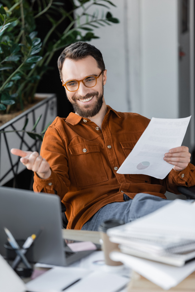 Радостный бородатый менеджер в стильных очках и футболках, держащий документ и указывающий на ноутбук во время видеоконференции возле размытых документов на рабочем столе в офисе - Фото, изображение