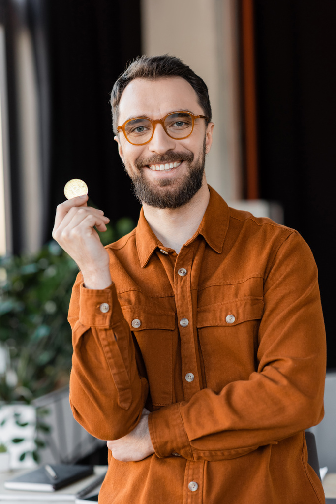 KYIV, UKRAINE - OCTOBER 18, 2022: charismatic and bearded businessman in trendy eyeglasses and shirt holding golden bitcoin and smiling at camera in modern office on blurred background - Photo, Image