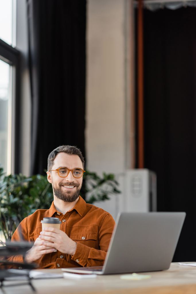 homem de negócios barbudo feliz em óculos elegantes e camisa segurando café para ir enquanto sentado no local de trabalho e olhando para laptop no escritório moderno em fundo borrado - Foto, Imagem