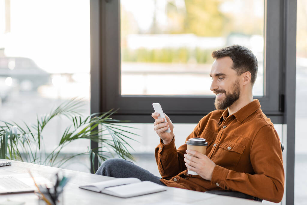 boldog és sikeres szakállas üzletember elvitelre ital üzenetküldés mobiltelefonon közel notebook, laptop, dekoratív növények és nagy ablakok az irodában - Fotó, kép