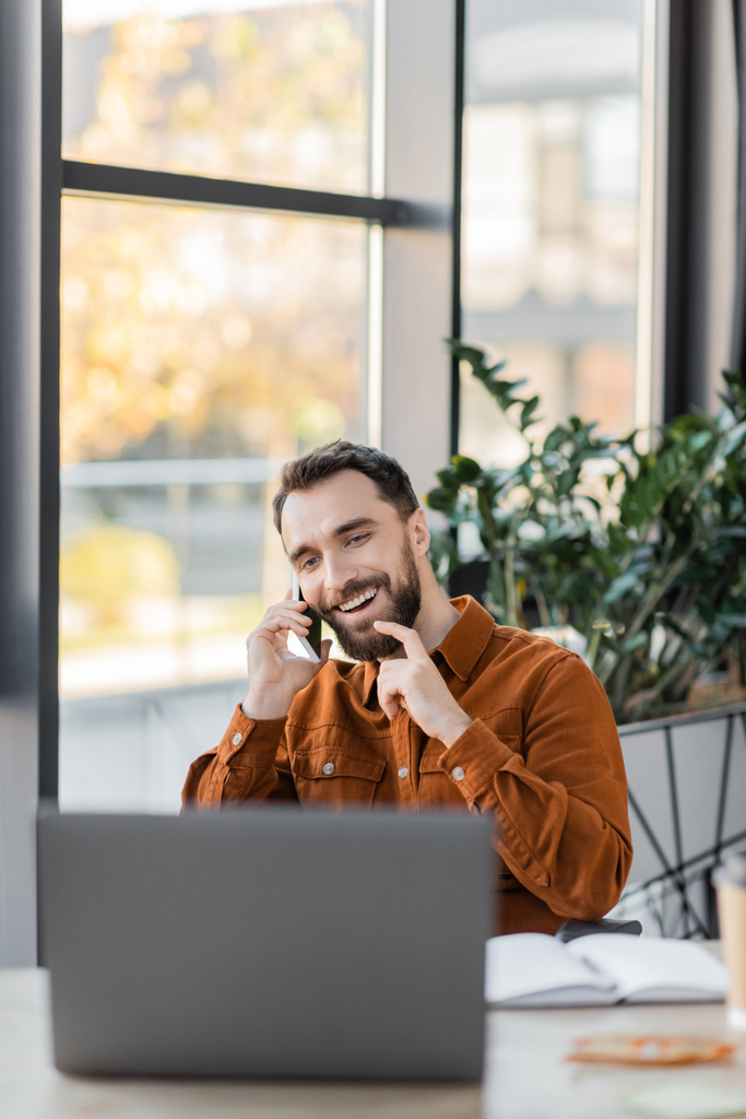 glimlachende zakenman aanraken baard en praten op mobiele telefoon in de buurt van notebook en wazig laptop naast decoratieve planten op de achtergrond in het hedendaagse kantoor - Foto, afbeelding