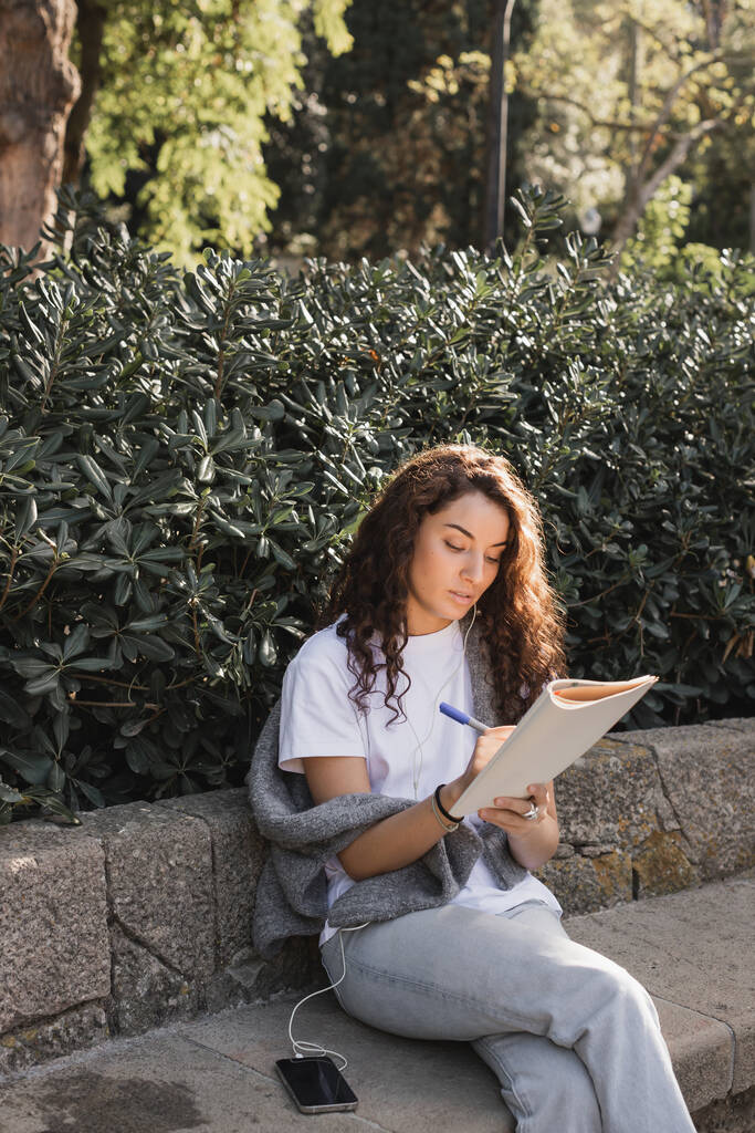 Junge und lockige brünette Frau schreibt am Notizbuch, während sie tagsüber im Park in Barcelona Musik in kabelgebundenen Kopfhörern in der Nähe des Smartphones auf einer steinernen Bank und grünen Büschen hört  - Foto, Bild
