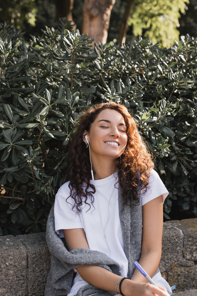Giovane donna bruna spensierata con gli occhi chiusi in abiti casual ascoltare musica in cuffia cablata e sorridente, tenendo il pennarello mentre si rilassa sulla panchina di pietra nel parco di Barcellona, Spagna  - Foto, immagini