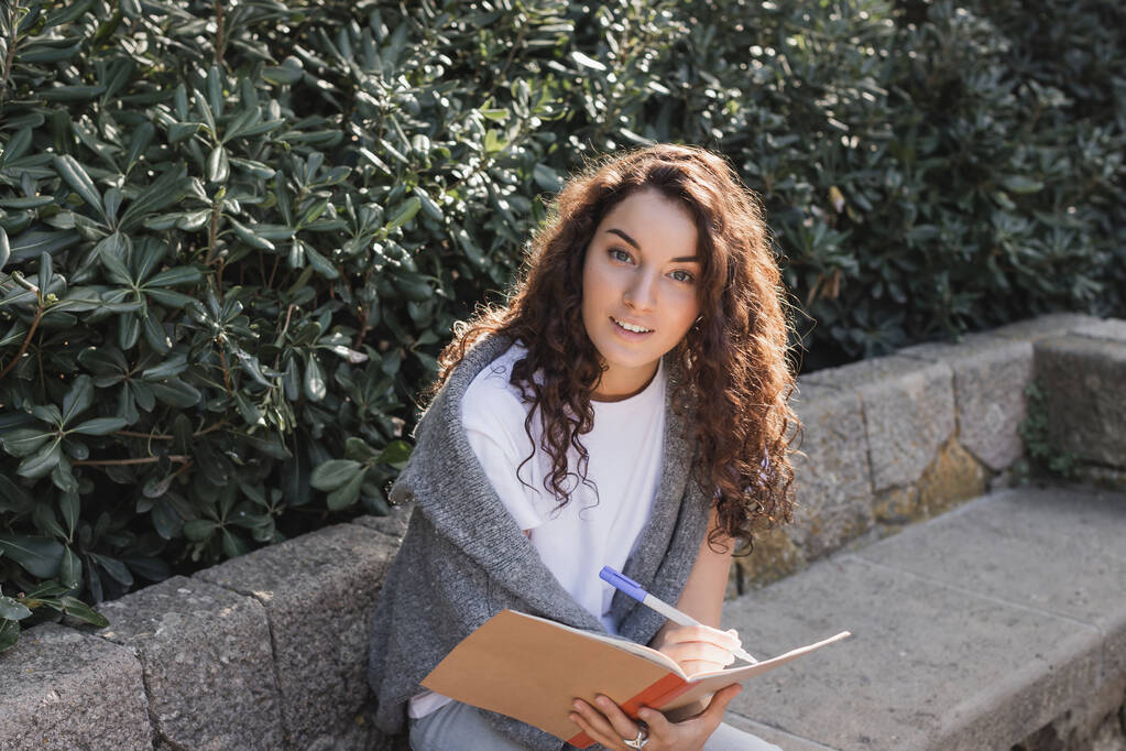 Unbekümmerte junge und lockige Frau in Pullover und T-Shirt, die in die Kamera schaut, während sie am Notizbuch schreibt und Zeit auf einer Bank in der Nähe grüner Büsche im Park in Barcelona, Spanien verbringt  - Foto, Bild