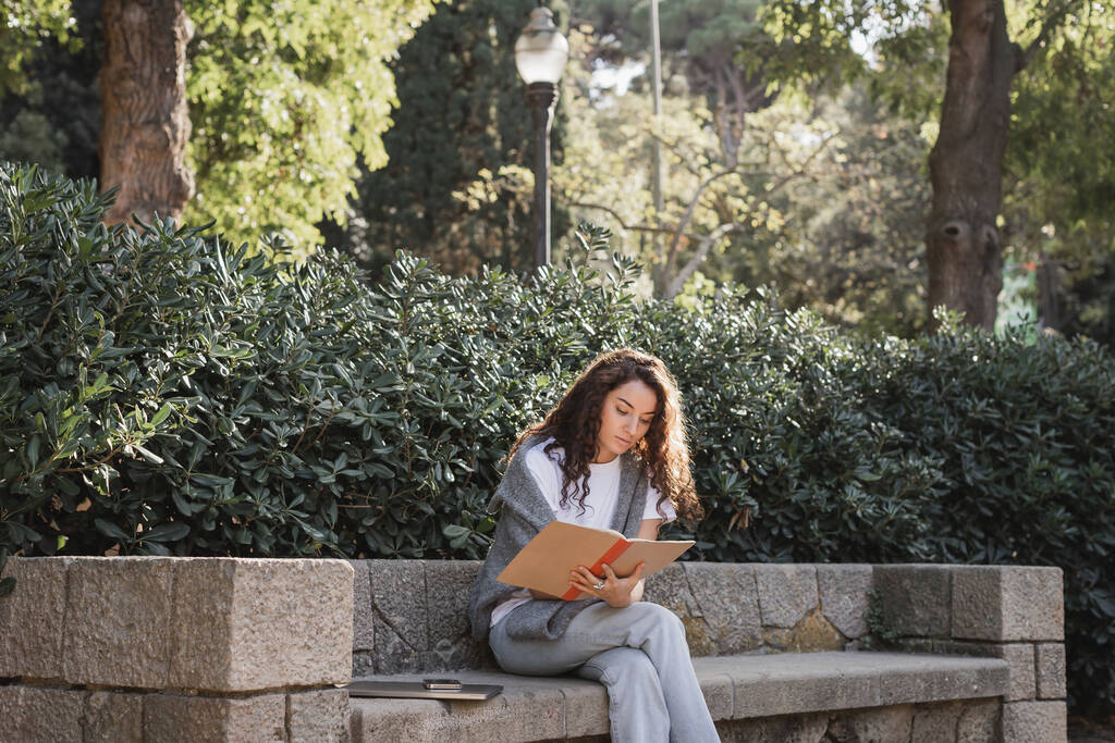 Młoda kręcona kobieta w luźnych ciuchach, trzymająca notatnik, siedząc w pobliżu urządzeń na kamiennej ławce i zielonych krzakach i spędzając czas w parku w dzień w Barcelonie, Hiszpania  - Zdjęcie, obraz