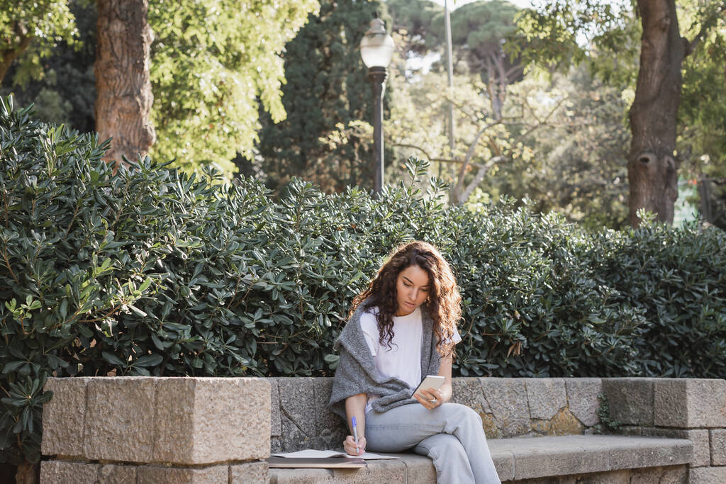 Young and curly freelancer in casual clothes using smartphone and writing on notebook while sitting near laptop on stone bench and green bushes in park in Barcelona, Spain  - Photo, Image