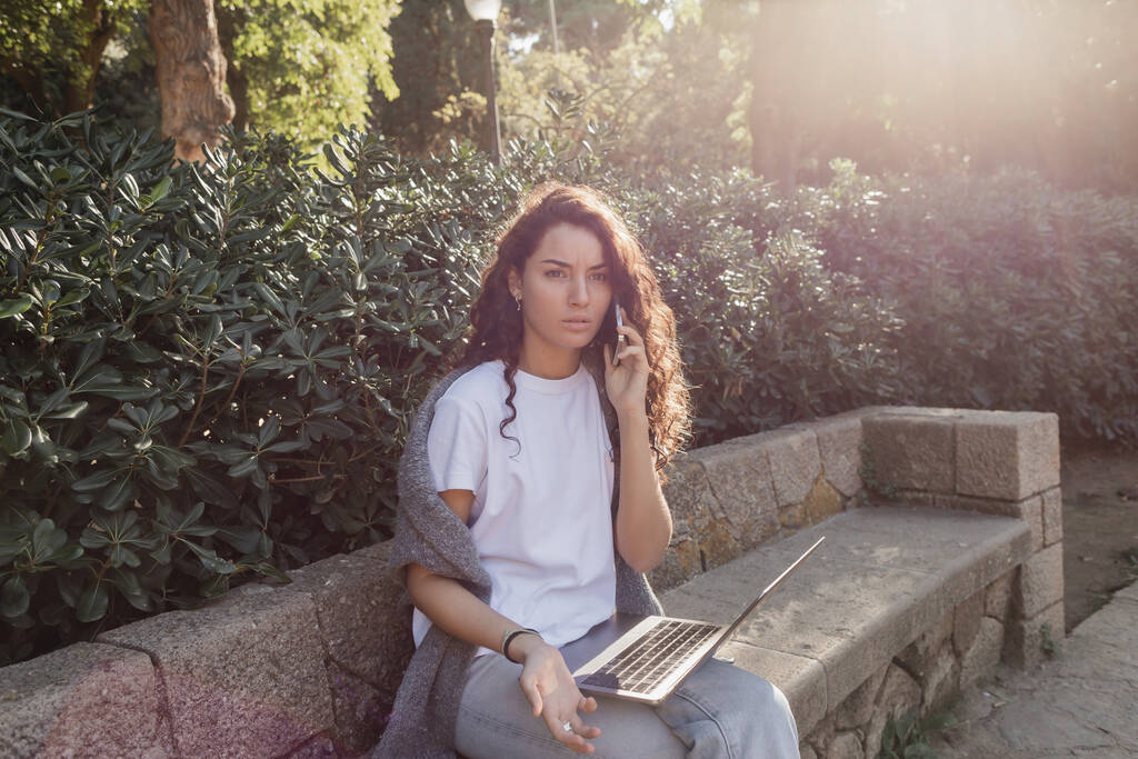 Pensive young and curly freelancer in casual clothes talking on smartphone near laptop while sitting on stone bench and spending time near green plants in park in Barcelona, Spain  - Photo, Image