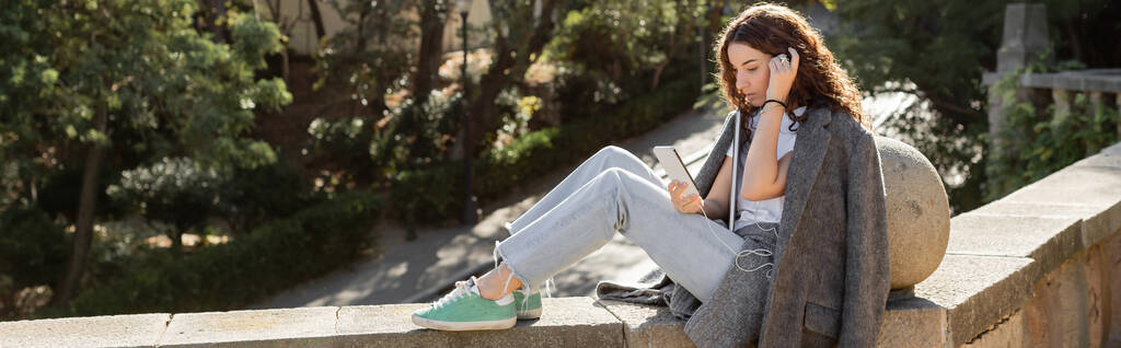 Jeune freelance bouclé en veste chaude à l'aide d'un smartphone avec écouteurs et tenant un ordinateur portable assis sur un banc en pierre dans un parc flou à Barcelone, Espagne, bannière  - Photo, image
