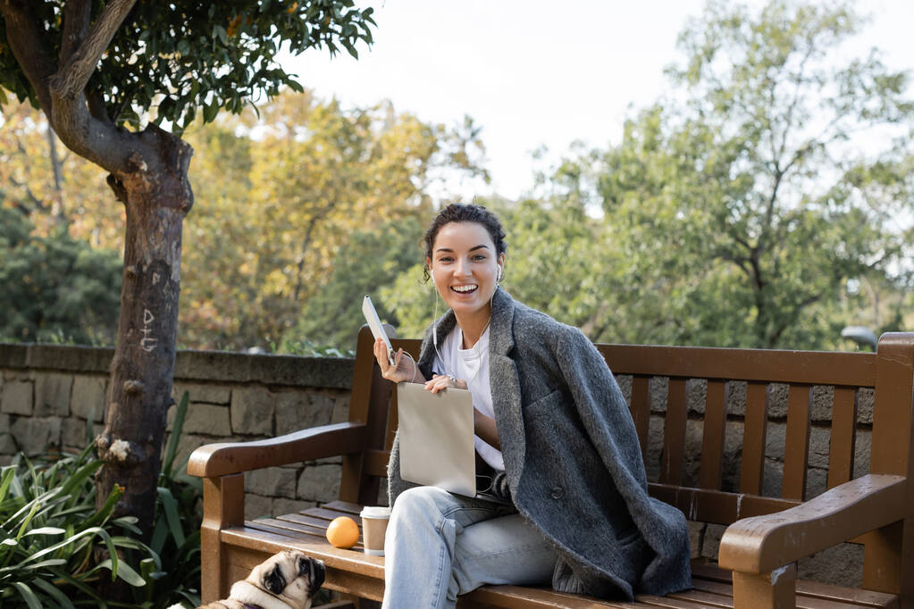 Cheerful young freelancer in warm jacket listening music in earphones, looking at camera and using devices near pug dog and takeaway coffee on wooden bench in park in Barcelona, Spain  - Photo, Image