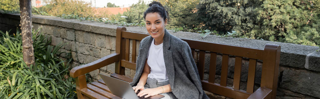 Wesoła młoda brunetka freelancer w ciepłej kurtce patrząc w kamerę podczas pracy na laptopie i siedząc na drewnianej ławce w parku w dzień w Barcelonie, Hiszpania, baner  - Zdjęcie, obraz