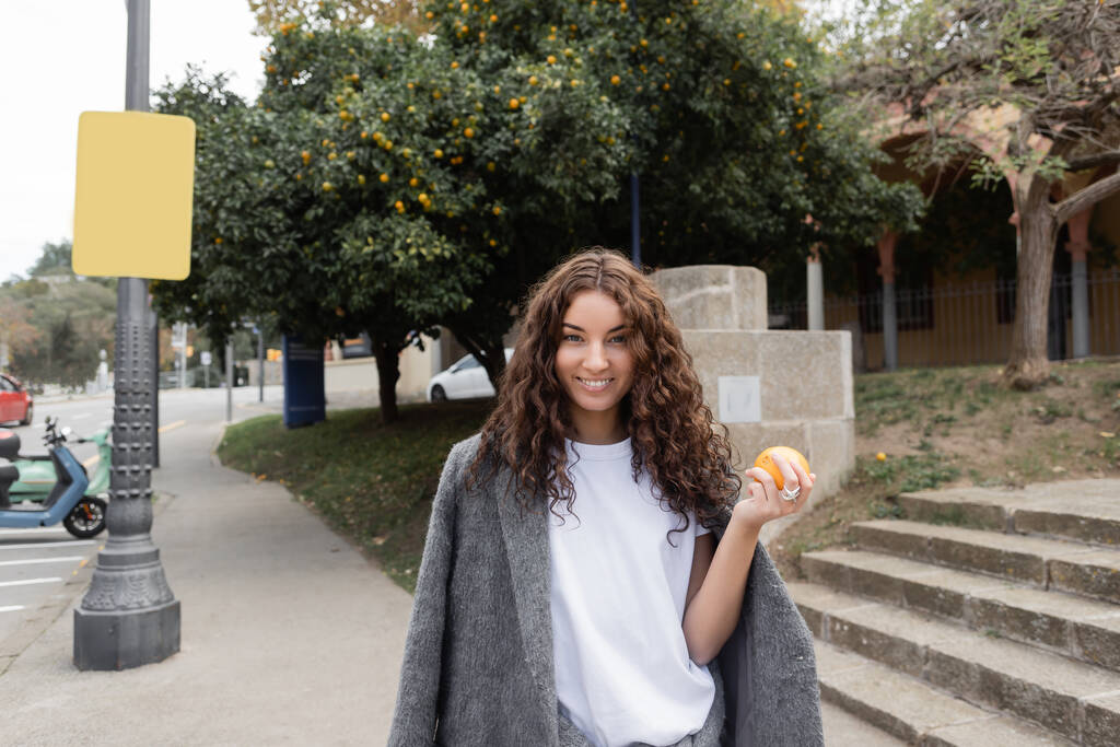 Sorridente giovane e riccia donna in giacca calda tenendo fresco arancione e guardando la fotocamera mentre in piedi su strada urbana offuscata sullo sfondo a Barcellona, Spagna, lampione, motorino - Foto, immagini