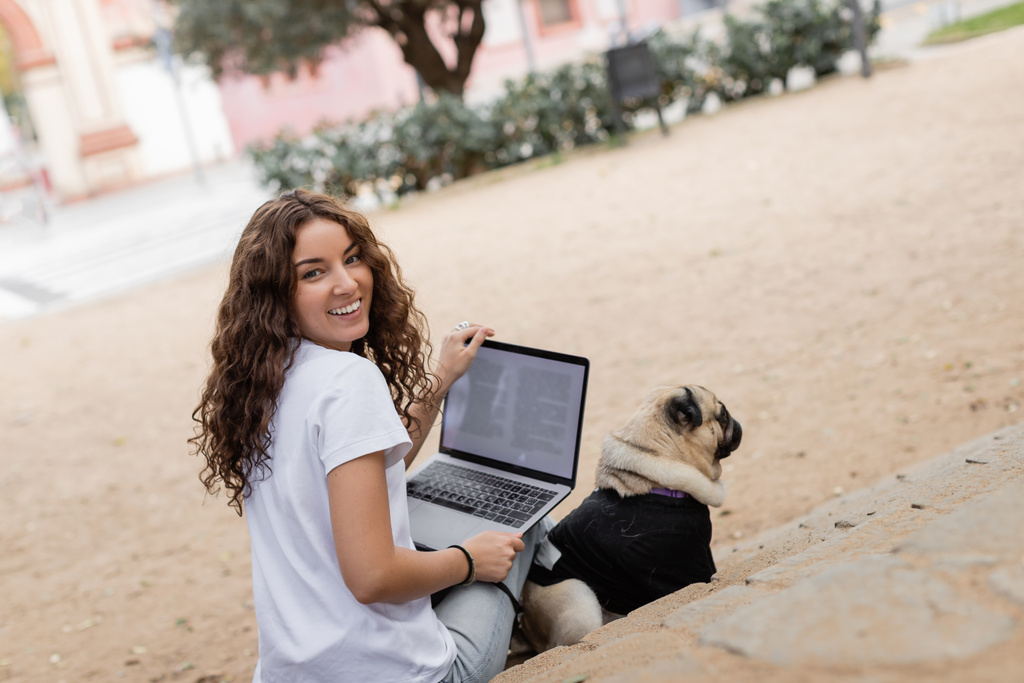 Веселая молодая кудрявая женщина в повседневной одежде смотрит в камеру, держа ноутбук рядом с собакой-мопсом на лестнице в размытом парке Барселоны, Испания, белая футболка  - Фото, изображение