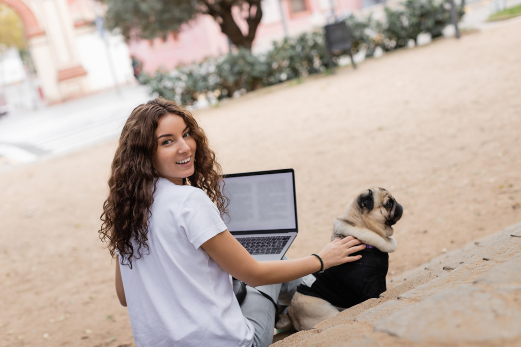 Pozytywny młody i kręcony wolny strzelec w luźnych ciuchach patrzący w kamerę trzymający laptopa i pieszczącego psa mopsa na schodach w zamglonym parku w Barcelonie, Hiszpania, biały t-shirt  - Zdjęcie, obraz