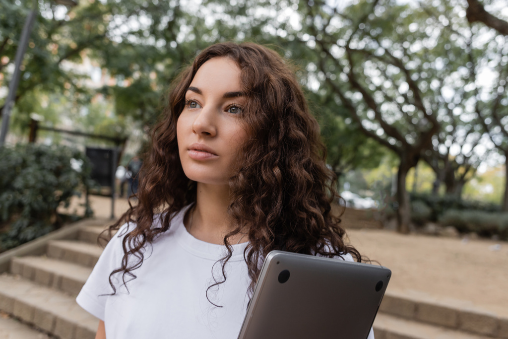 Ritratto di giovane donna bruna in t-shirt bianca che tiene il computer portatile e distoglie lo sguardo mentre trascorre del tempo nel parco sfocato di Barcellona, Spagna  - Foto, immagini