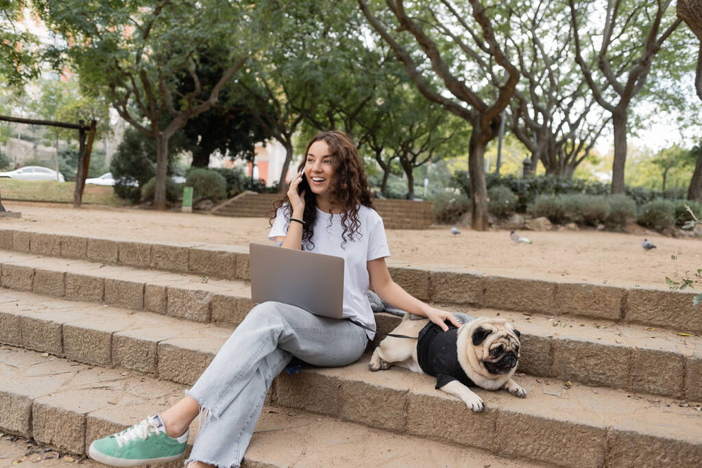 Cheerful brunette freelancer in casual clothes talking on smartphone near laptop and petting pug dog on stairs while spending time in blurred park in Barcelona, Spain  - Photo, Image