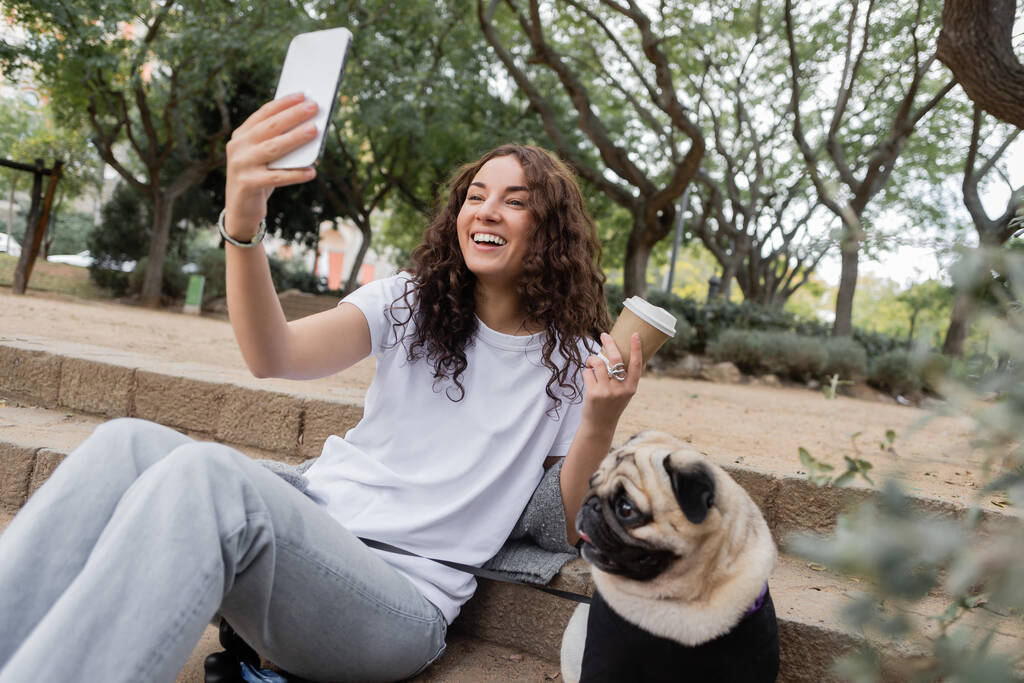 Mujer alegre joven y rizada en ropa casual que tiene videollamada en el teléfono inteligente y la celebración de un café para llevar cerca del perro pug sentado en las escaleras en el parque borroso durante el día en Barcelona, España  - Foto, Imagen
