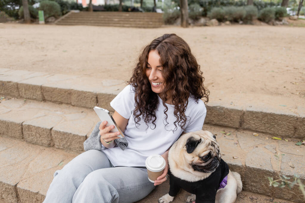 Overjoyed young curly woman in casual clothes using smartphone and holding coffee to go while sitting near pug dog on stairs in blurred park in Barcelona, Spain  - Photo, Image