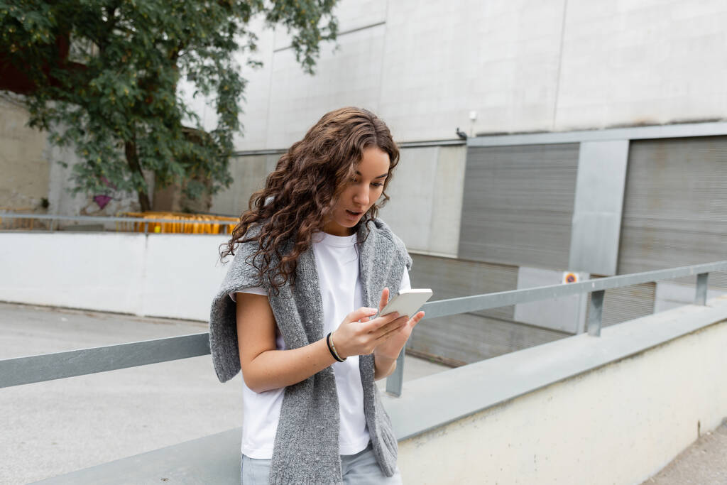 Junge lockige Frau in warmem Pullover auf den Schultern mit Smartphone, während sie tagsüber in Barcelona auf einer Straße mit Gebäuden im Hintergrund steht  - Foto, Bild
