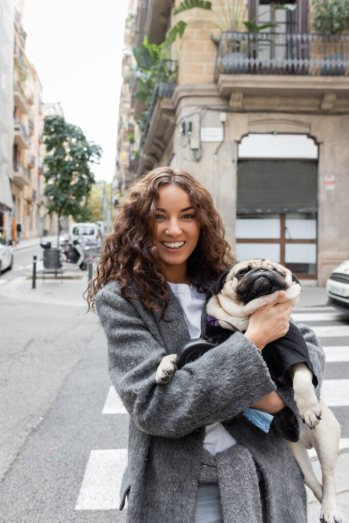 Cheerful young woman in warm jacket looking at camera and holding pug dog on hands while standing near blurred building on urban street at daytime in Barcelona, Spain  - Photo, Image