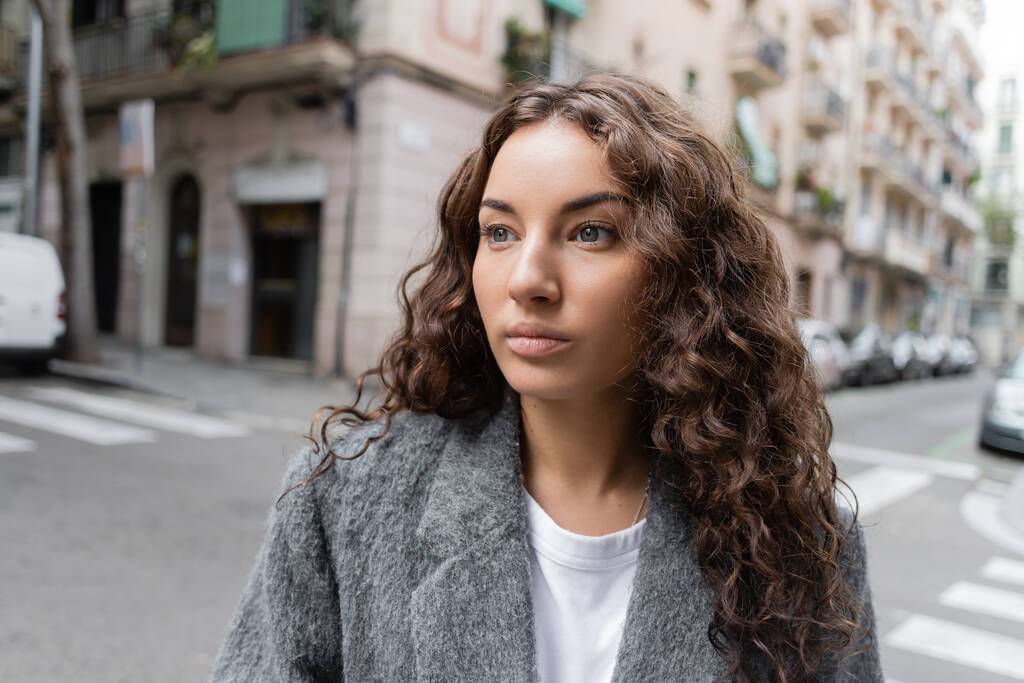 Brunette and pretty young woman in casual jacket looking away while standing on blurred city street with buildings at background in Barcelona, Spain  - Photo, Image