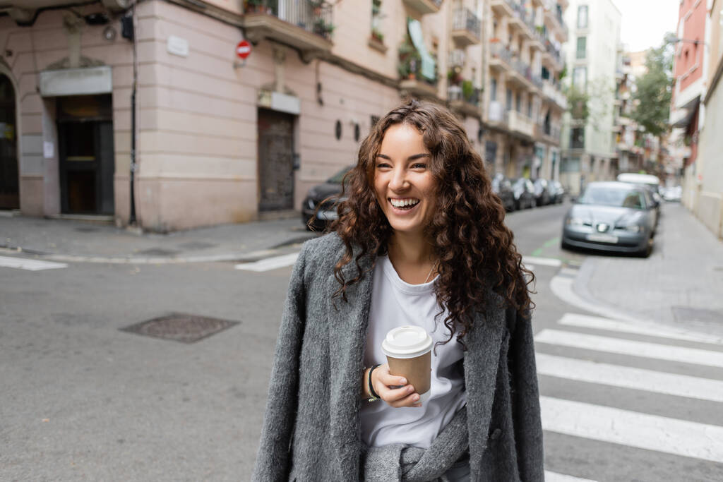 Donna giovane e riccia positiva in giacca casual che tiene il caffè in tazza di carta e distoglie lo sguardo sulla strada della città offuscata con auto ed edifici durante il giorno a Barcellona, Spagna  - Foto, immagini