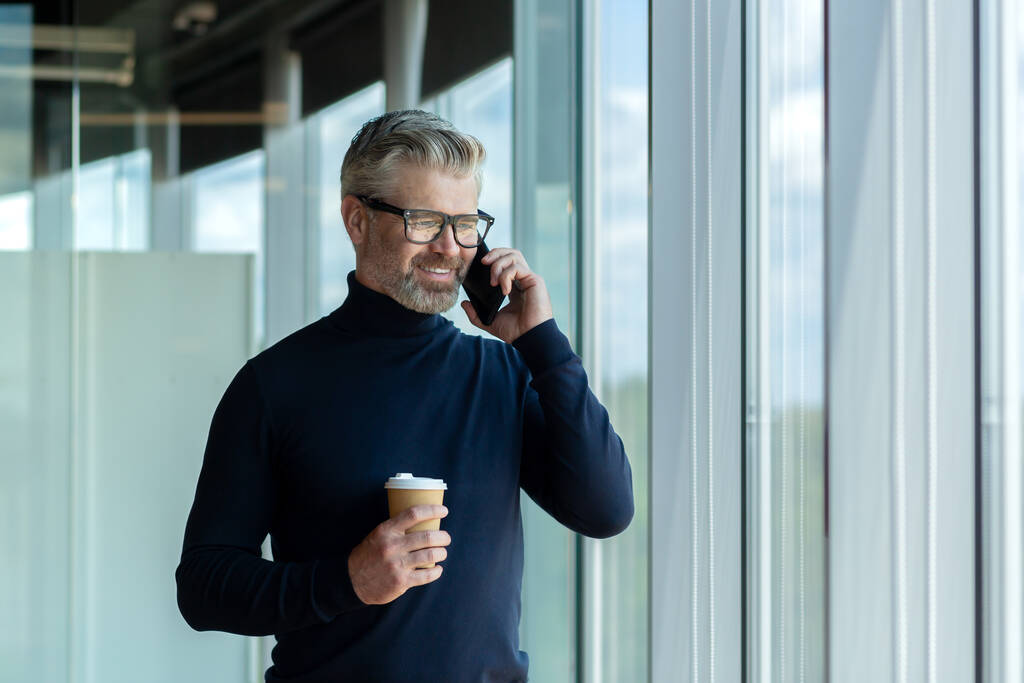 Przystojny starszy biznesmen stojący w biurze przy oknie z filiżanką kawy i uśmiechnięty rozmawiający przez telefon.. - Zdjęcie, obraz