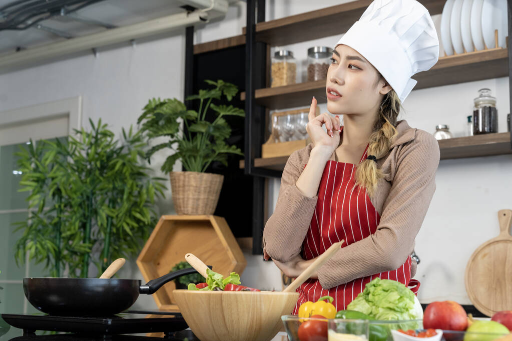 ポートレートアジアの美しい女性シェフの帽子とエプロンポーズは、キッチンで調理している間、カウンターの電気ストーブ、卵、野菜、キッチン機器の場所でのパン、コピースペース - 写真・画像