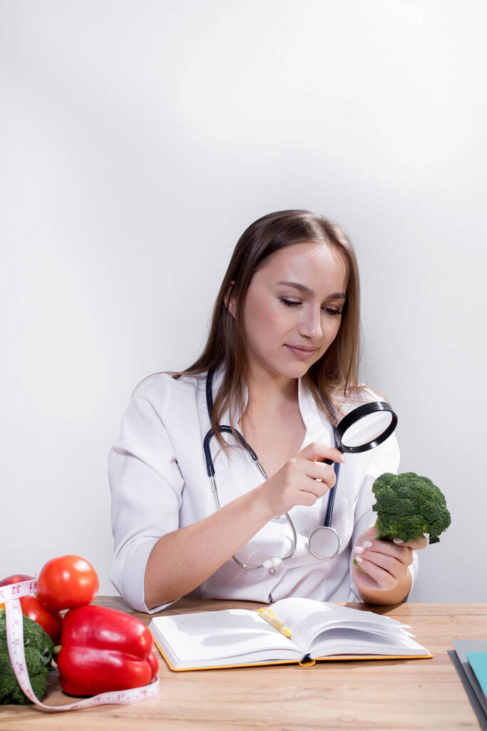 A female nutritionist looks through a magnifying glass at broccoli in her office. Healthy Eating. - Φωτογραφία, εικόνα