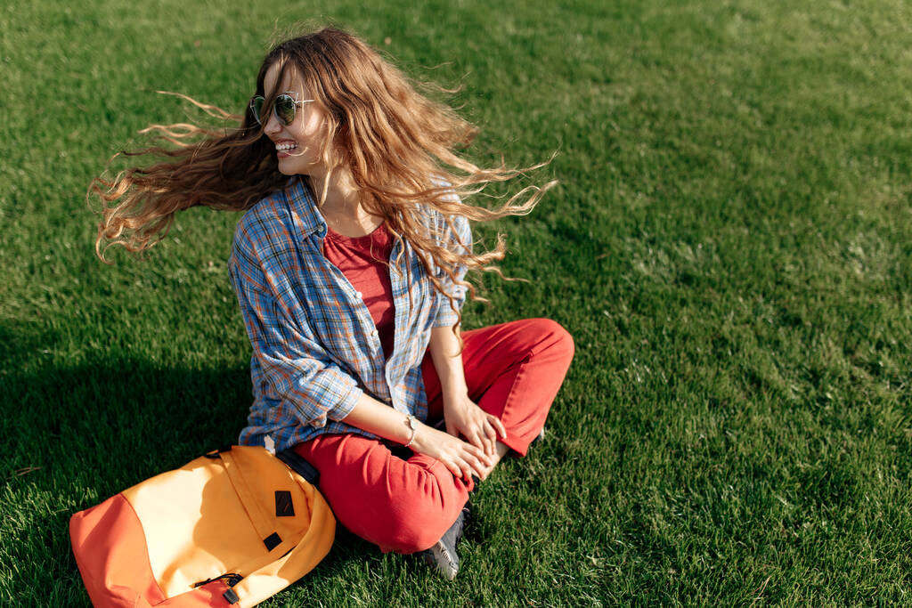 Glückliche junge blonde Hipster-Studentin mit Rucksack sitzt auf Gras an der Universität - Foto, Bild