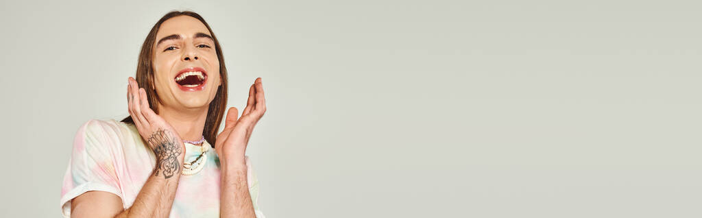tatuado e feliz jovem gay homem com longo cabelo olhando para câmera com boca aberta e gesticulando com as mãos no fundo cinza, orgulho mês conceito, banner  - Foto, Imagem