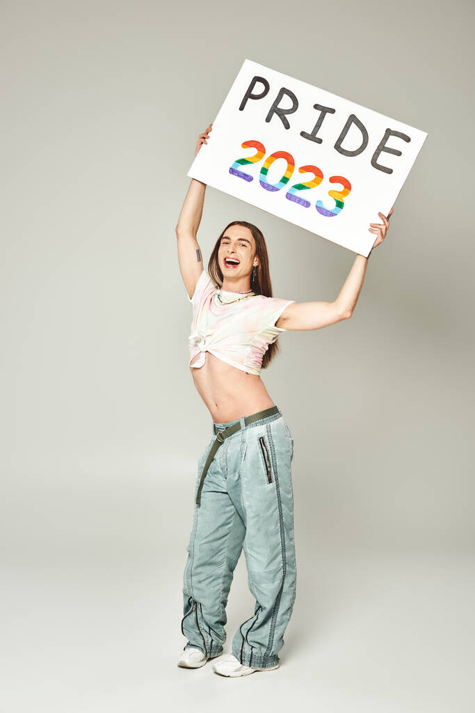 cheerful young gay man with tattoo and long hair standing in tied knot on t-shirt showing his belly and denim jeans while holding pride 2023 placard on grey background  - Photo, Image