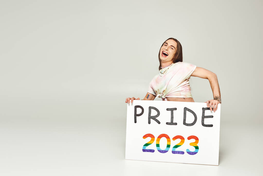 возбужденный молодой гей-активист с татуировкой и длинными волосами стоя с открытым ртом и держа гордость 2023 плакат на сером фоне, lgbt сообщества праздник в июне  - Фото, изображение
