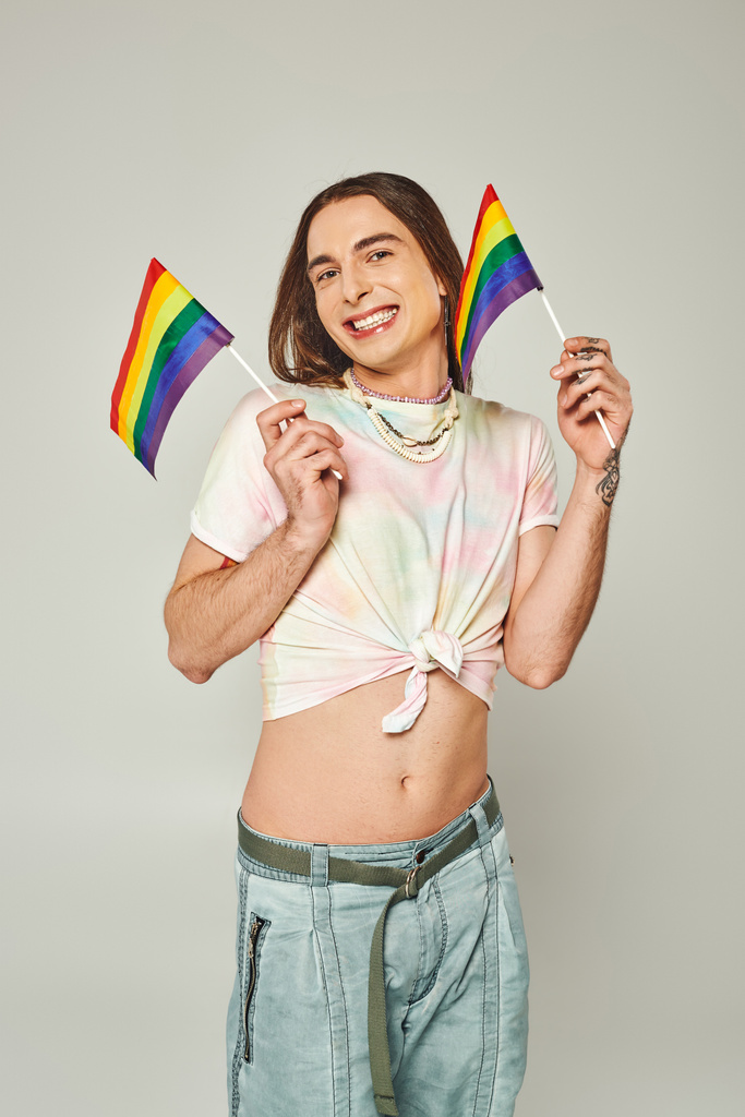 счастливый и татуированный гей с длинными волосами и голым животом, стоящий в джинсах в джинсах, держа в руках радужные флаги для гордости месяц на сером фоне  - Фото, изображение