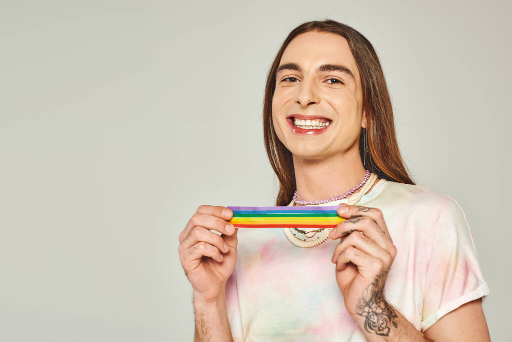 happy and tattooed gay man with long hair and tie dye t-shirt holding rainbow lgbt flag for pride month and smiling while looking at camera on grey background  - Photo, Image
