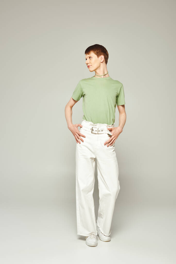 volledige lengte van de jonge queer persoon met glanzende lip gloss staan in groen t-shirt en witte spijkerbroek terwijl weg te kijken tijdens trots maand op grijze achtergrond - Foto, afbeelding