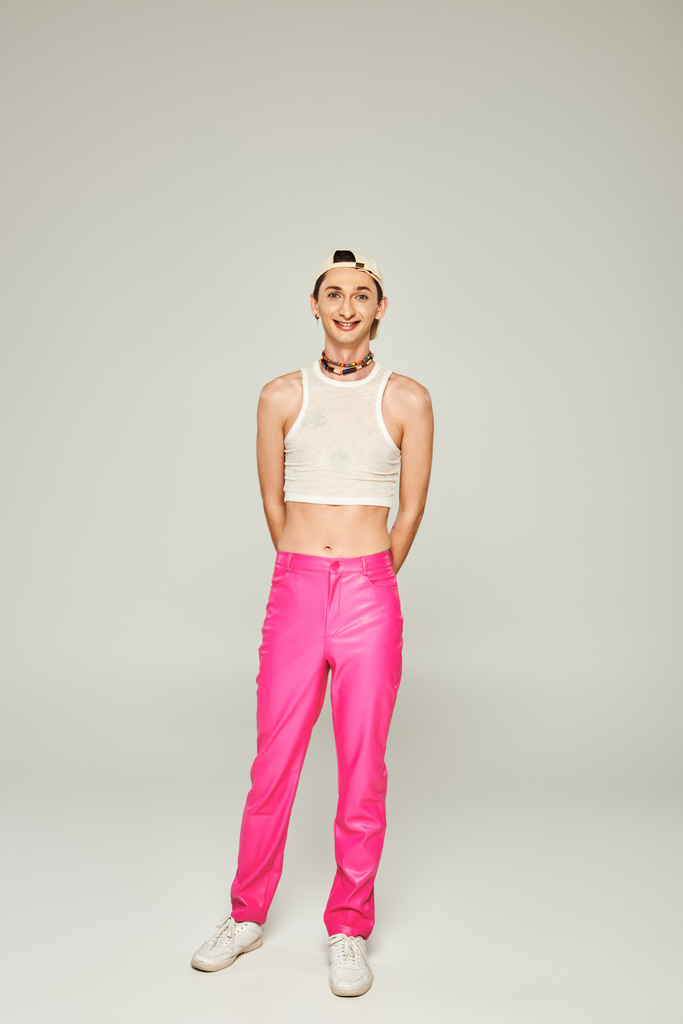 volledige lengte van gelukkig en getatoeëerd gay man in baseball cap, gewas top en roze broek glimlachen terwijl poseren op grijze achtergrond, trots dag concept  - Foto, afbeelding