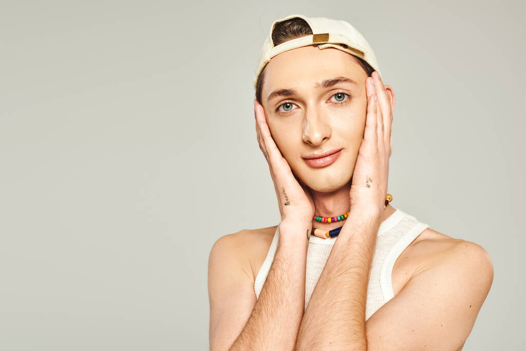 portrait of tattooed young gay man with blue eyes posing in baseball cap and colorful beads and looking at camera on grey background, pride day concept  - Photo, Image