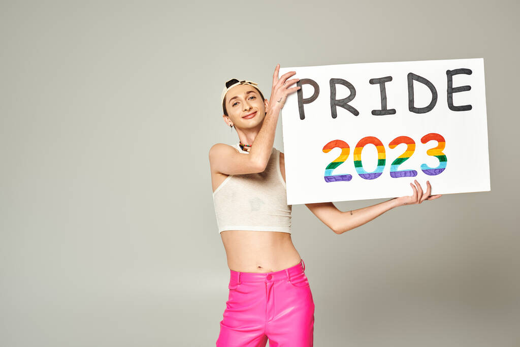 татуйований і задоволений молодий гей чоловік у бейсбольній шапці, верхній частині врожаю, і рожеві штани, що тримають гордість плакату 2023 року, стоячи і дивлячись на камеру на сірому фоні
  - Фото, зображення