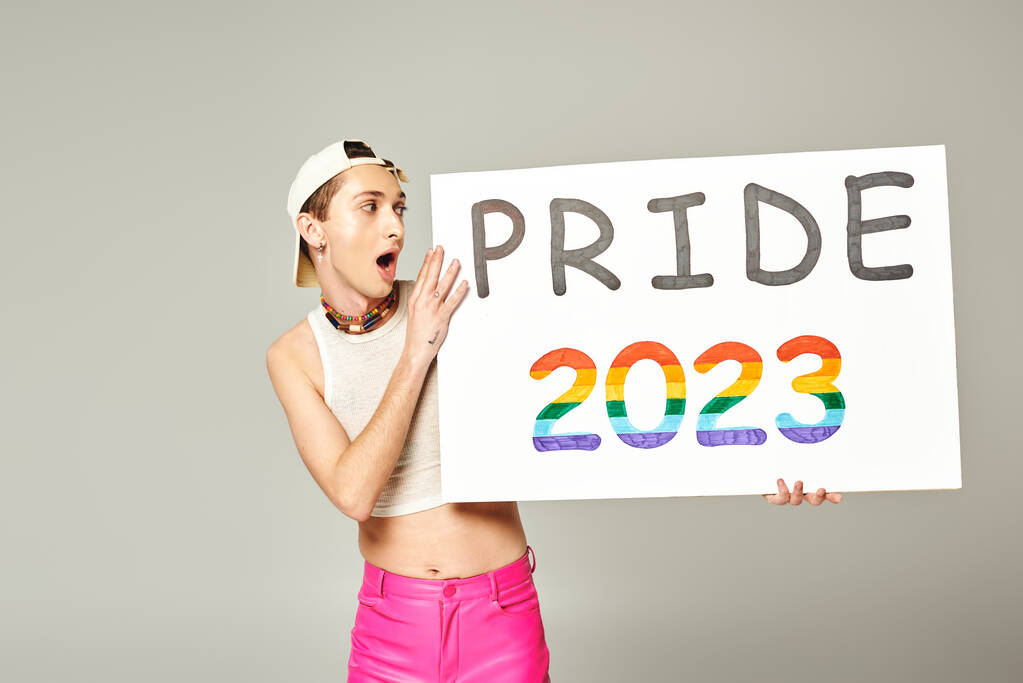 tattooed and shocked gay man in baseball cap, crop top, and pink pants holding pride 2023 placard while standing with opened mouth on grey background  - Photo, Image