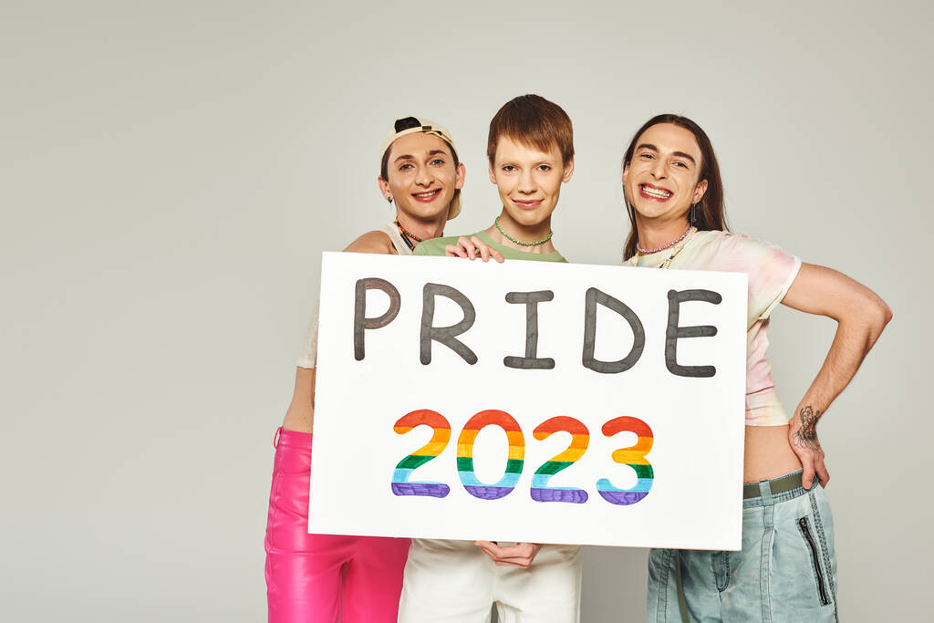 változatos és fiatal lgbtq közösségi barátok tetovált áll színes ruhák és gazdaság büszkeség 2023 placard, miközben a kamera a stúdióban, szürke háttér  - Fotó, kép