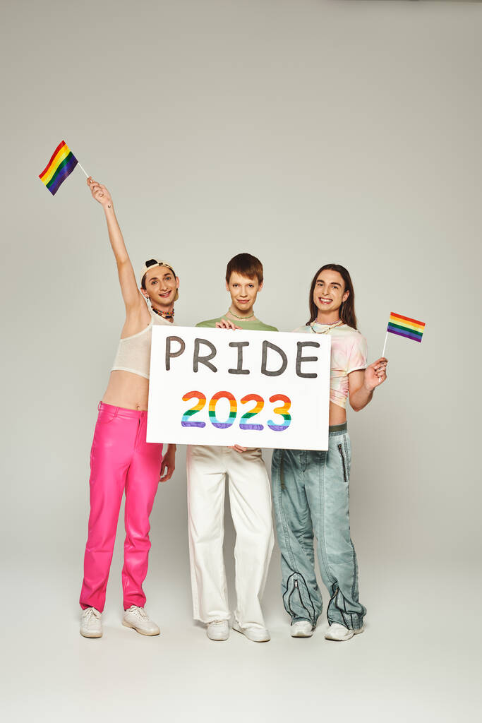 gai et tatoué gay les hommes dans coloré vêtements debout avec arc-en-ciel drapeaux près queer ami tenant fierté 2023 pancarte tout en célébrant lgbt vacances communautaires, fond gris, studio  - Photo, image