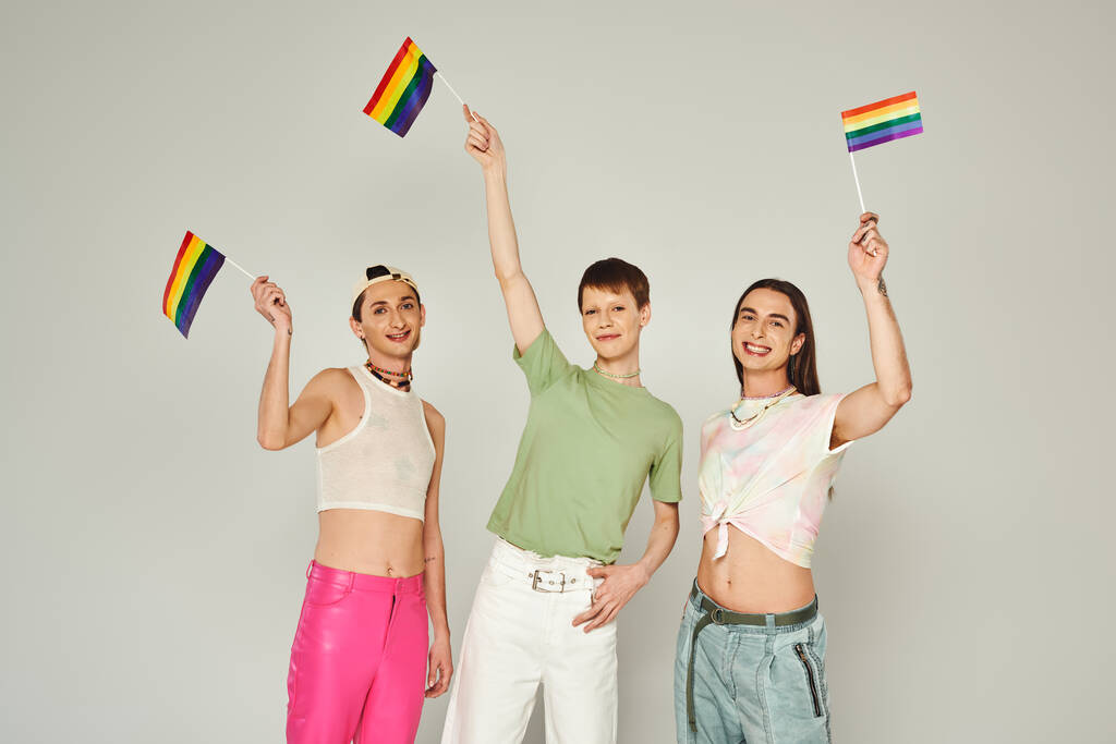 felizes e jovens amigos da comunidade lgbtq com tatuagens em pé em roupas coloridas e segurando bandeiras do arco-íris enquanto olha para a câmera em estúdio, fundo cinza  - Foto, Imagem