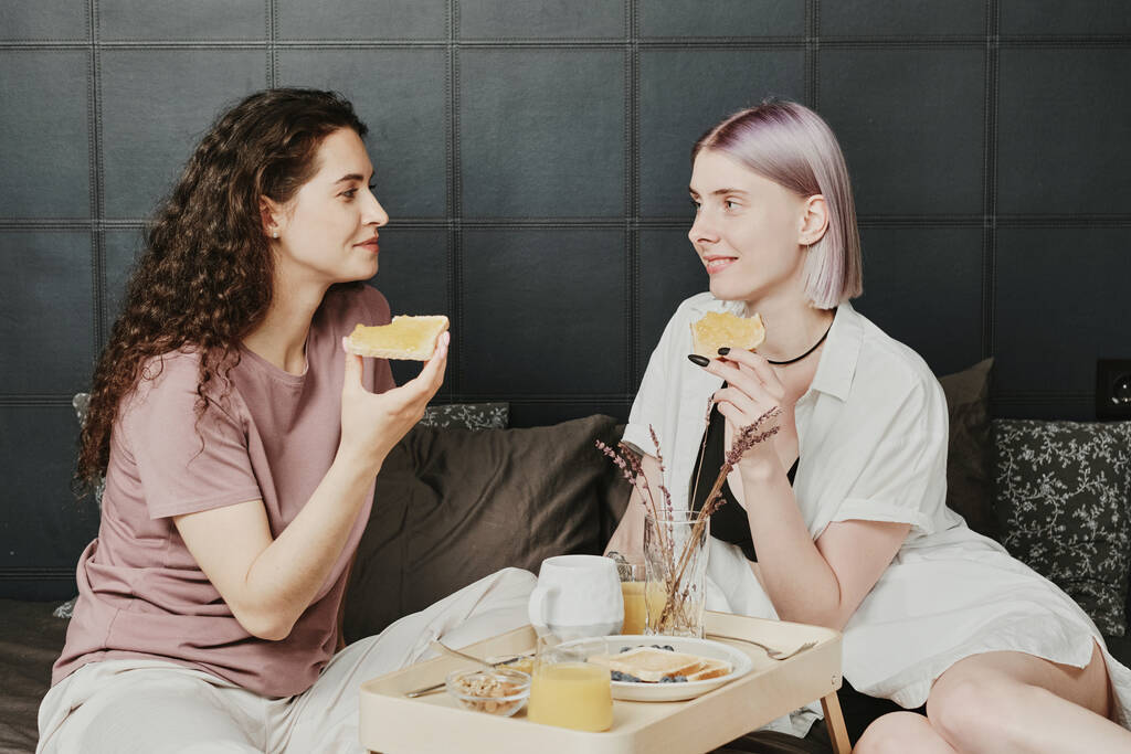 pozytywny młody biały Lesbijki para siedzi na łóżko z taca i jedzenie toasty w rano - Zdjęcie, obraz