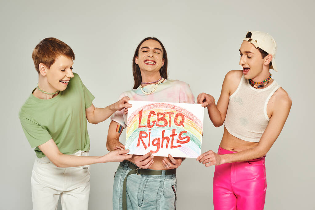 happy lgbt friends in colorful clothes holding placard with lgbtq rights lettering while standing together and smiling on pride month, grey background  - Photo, Image