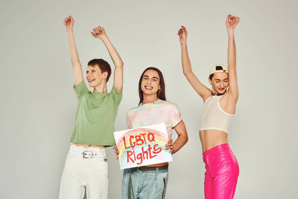 fröhliche lgbt-Freunde in bunten Kleidern tanzen mit erhobenen Händen neben Aktivisten mit Plakat mit lgbtq-Schriftzug und lächeln auf stolzem Monat, grauem Hintergrund  - Foto, Bild