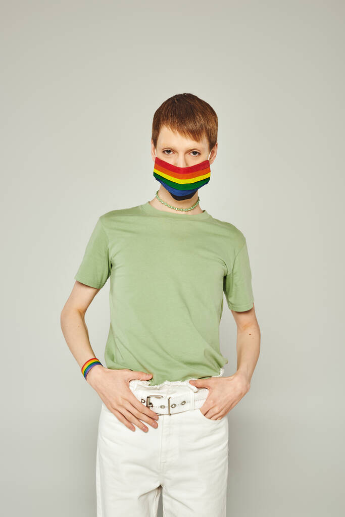 portrét mladého teplouše stojící v zeleném tričku a vlajkové masce lgbt při pohledu na kameru a pózování během měsíce pýchy na šedém pozadí - Fotografie, Obrázek