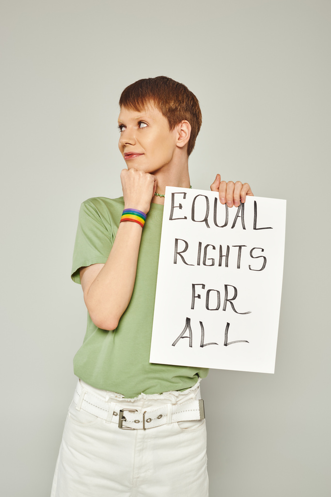 portrait of smiling queer activist wearing lgbt flag bracelet and holding placard with equal rights for all lettering looking away during pride month on grey background - Photo, Image